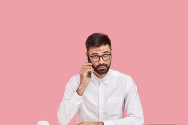 Serious unshaven intelligent man calls customer service, has telephone conversation in break of work, dressed in white shirt, focused upwards, wears transparent spectacles, surrounded with devices