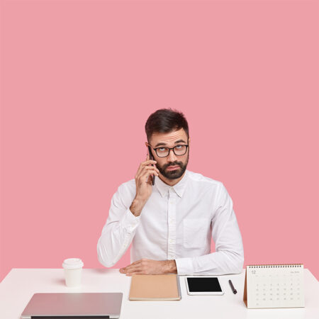 Serious unshaven intelligent man calls customer service, has telephone conversation in break of work, dressed in white shirt, focused upwards, wears transparent spectacles, surrounded with devices